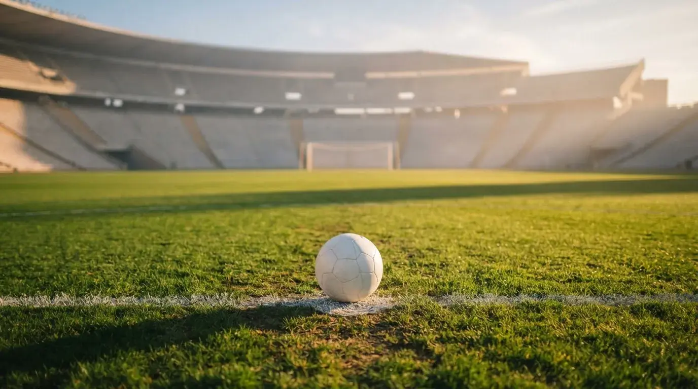 Ein Fußball liegt auf dem Elfmeterpunkt eines leeren Rasenplatzes, im Hintergrund eine Tribüne im Dunst.