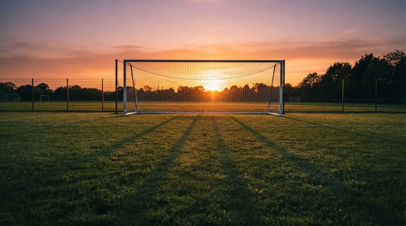 Ein Fußballtor mit Netz bei Sonnenuntergang auf einem leeren Platz, die Schatten der Torpfosten liegen lang auf dem Gras.