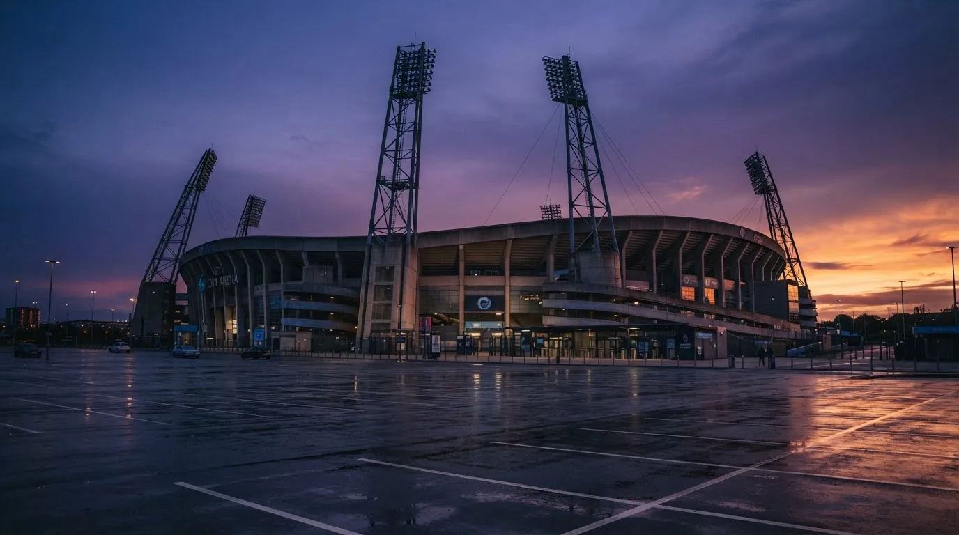 Ein Fußballstadion von außen in der Dämmerung, hohe Flutlichtmasten vor einem violetten Himmel, davor ein leerer Parkplatz.