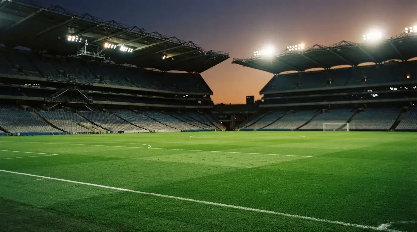 Eine leere Stadiontribüne unter Flutlicht aus der Ferne betrachtet, die Rasenfläche leuchtet grün in der Dämmerung.