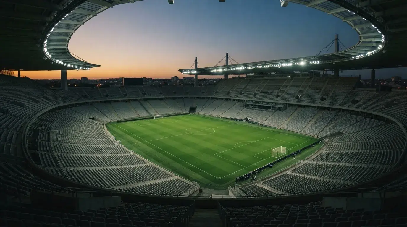 Eine leere Stadiontribüne unter Flutlicht aus der Ferne betrachtet, die Rasenfläche leuchtet grün in der Dämmerung.