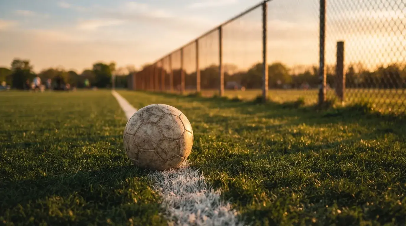 Ein Fußball liegt am Rand eines Rasens neben einer weißen Linie, im Hintergrund ein Zaun in der Abendsonne.