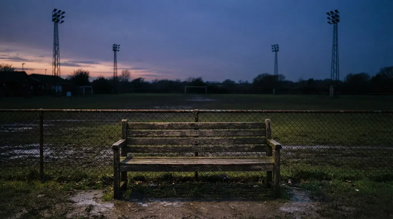 Eine leere Parkbank am Rand eines Fußballfelds in der Abenddämmerung, im Hintergrund ragen dunkle Flutlichtmasten in den Himmel.
